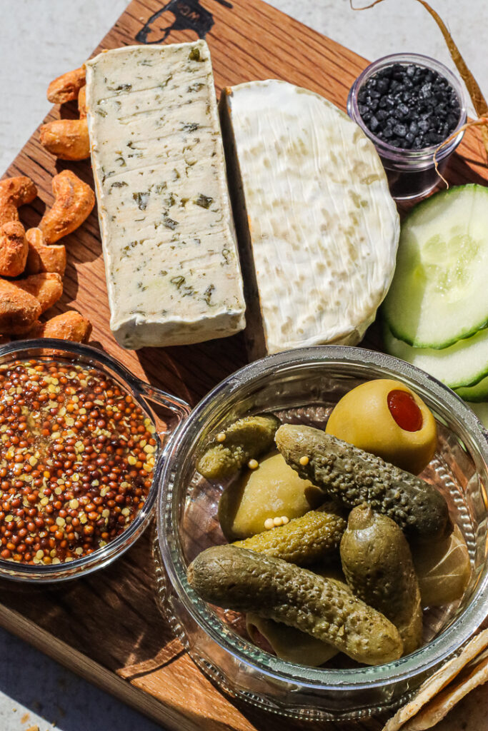 cut cheese wheel with truffle specs, cornichons, mustard, cucumbers, and nuts on a cheese board.