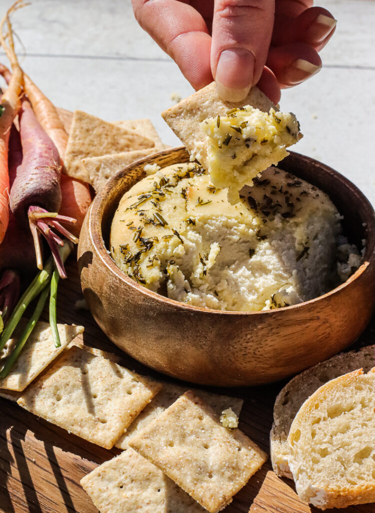 hand dipping a cracker into soft, herb topped cheese.
